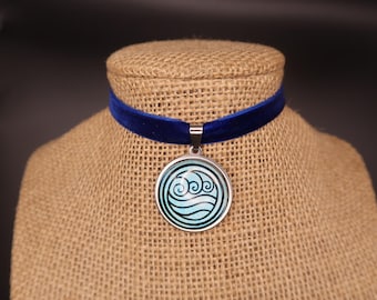 May include: A blue velvet choker necklace featuring a circular silver-toned pendant. The pendant displays a light blue background with a dark blue graphic design of stylized swirling water waves centered within the circular frame.