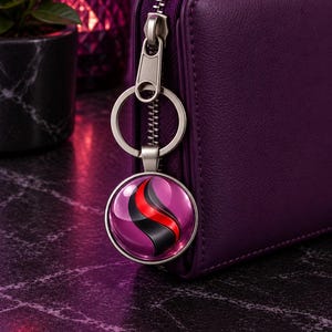 May include: A deep purple zippered wallet featuring a circular metal keychain attached to the zipper pull. The keychain displays a glossy, rounded cabochon with a stylized graphic design consisting of a black and red curved stripe on a purple background.
