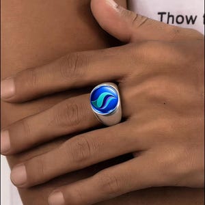 May include: A silver signet ring featuring a circular inlay with a glossy blue background and a stylized, swirling teal and purple graphic design. The ring has a smooth, polished metallic band that sits securely on the finger.