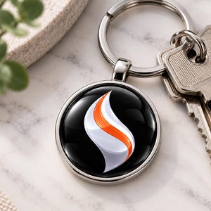 Op de afbeelding: Een ronde metalen sleutelhanger met een glanzend zwarte inzet met een gestileerde witte en oranje vlamvormige afbeelding. De hanger is bevestigd aan een standaard zilverkleurige sleutelring, waar ook een set metalen sleutels aan hangt.