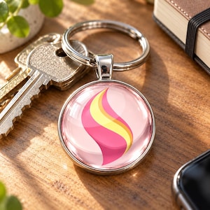 Könnte beinhalten: Ein runder Metallschlüsselanhänger mit einer glänzenden, gewölbten Oberfläche und einem stilisierten Grafikdesign. Das Motiv besteht aus einer zentralen, geschwungenen, flammenartigen Form in leuchtendem Pink und Gelb auf hellrosa Grund.