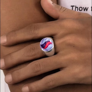 May include: A silver-toned ring featuring a circular cabochon inlay. The inlay displays a smooth, glossy surface with a central vertical red stripe flanked by two dark purple curved shapes against a light lavender and white background.