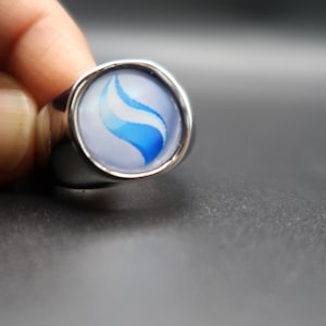 May include: A silver-toned metal ring featuring a circular cabochon inlay. The inlay displays a smooth, light blue background with a prominent, curved, darker blue flame-like graphic centered on the surface of the ring face.
