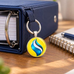 May include: A circular keychain featuring a glossy yellow background with a central stylized flame-like graphic in blue, white, and black. The charm is attached to a silver-toned metal split ring, which is secured to the spine of a dark blue binder.