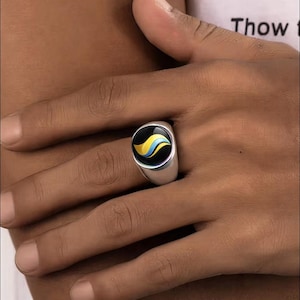 Puede incluir: Un anillo plateado con una cara negra que presenta un diseño de onda amarilla y azul. El anillo se muestra en el dedo de una persona. La palabra "Thow" es parcialmente visible en el fondo.