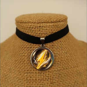 Puede incluir: Un collar de gargantilla de terciopelo negro con un colgante plateado. El colgante es un diseño circular con un rayo amarillo. El rayo tiene un efecto brillante, sobre un fondo oscuro.