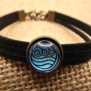 May include: Black suede bracelet with a round, bronze-colored charm. The charm features a blue and white design of swirling water. The bracelet has a double-strand design and a bronze clasp.