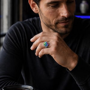 May include: A silver-toned ring featuring a circular face with a vibrant, iridescent, multi-colored inlay displaying a dark, stylized DNA double helix graphic. The ring has a polished metallic band and sits on a finger, showcasing the reflective surface.