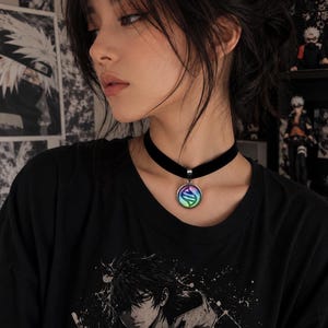 May include: A black velvet choker necklace featuring a circular pendant with a metallic silver frame and a colorful, iridescent gradient center displaying a stylized DNA-like helix symbol, worn against a black t-shirt with a graphic print.