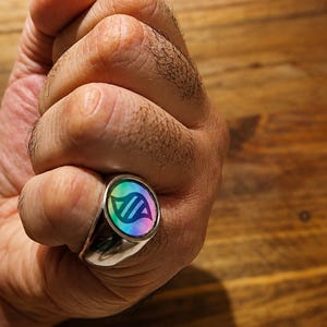 May include: A silver-toned metal ring featuring a circular face with a holographic, iridescent finish. The center of the ring displays a dark, stylized graphic symbol resembling a double helix or abstract geometric shape set against the rainbow colors.