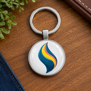 Könnte beinhalten: Ein runder Metallschlüsselanhänger mit einer glänzenden weißen Einlage, die eine stilisierte Flammengrafik zeigt. Das Flammendesign besteht aus einer großen dunkelblauen geschwungenen Form, einem gelben Mittelteil und einem dünnen rosa Rand.