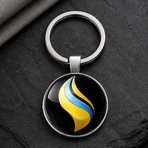 May include: A circular metal keychain featuring a glossy black insert with a stylized flame graphic in yellow and light blue. The design includes a silver-toned metal ring and a polished frame that holds the central circular emblem securely in place.