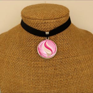 May include: A black velvet choker necklace featuring a round silver-toned pendant. The pendant contains a glossy, circular insert displaying a stylized pink and white swirl design with a central curved shape against a vibrant pink background.