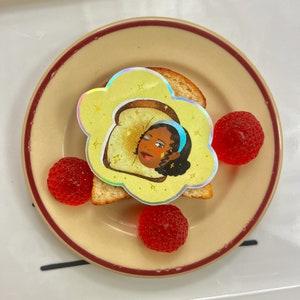 May include: A yellow holographic sticker featuring a cartoon illustration of a woman's face inside a slice of bread. The sticker is on a beige plate with red candy hearts.