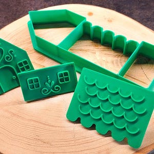 May include: Green plastic cookie cutters in the shape of a gingerbread house, including a house front with heart and window details, a roof, and walls. The cutters are arranged on a wooden surface.