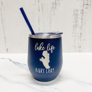 May include: A navy blue stainless steel tumbler with a white lid and a blue straw. The tumbler has the text "lake life" and "Burt Lake" printed on it in white.