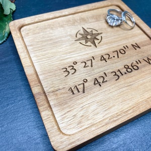 May include: A wooden ring dish with a compass rose design and coordinates engraved on the surface. The coordinates are 33° 27' 42.70" N 117° 42' 31.86" W.