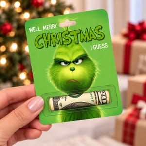 May include: A green rectangular card featuring a graphic of a furry green character with a grumpy expression. The text reads WELL, MERRY CHRISTMAS I GUESS. A clear plastic blister pack at the bottom holds a rolled-up twenty-dollar bill.