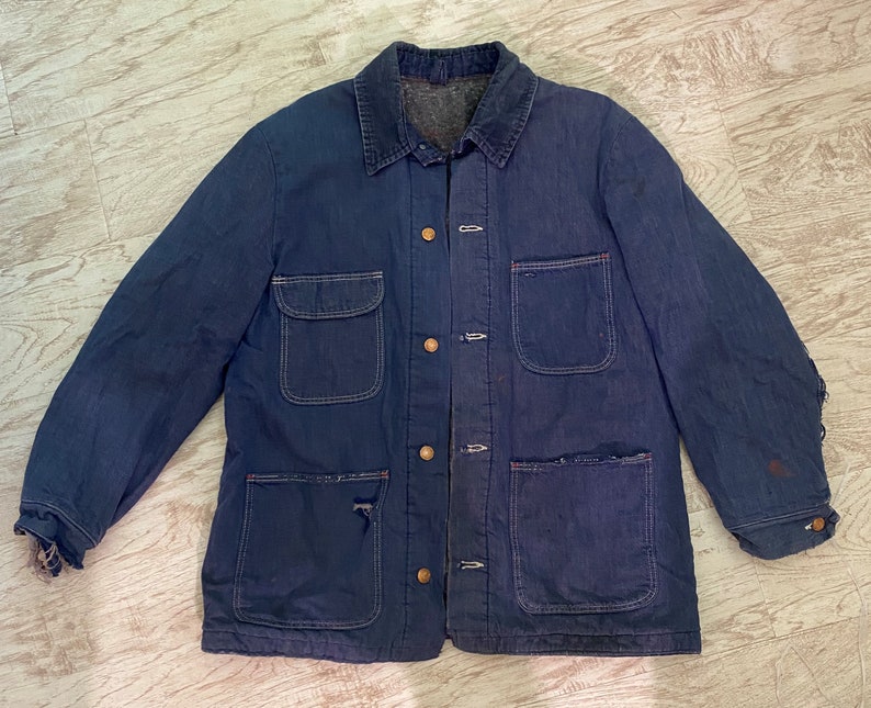May include: A dark blue denim work jacket with multiple pockets and a button closure. The jacket has a worn and faded look, with some visible wear and tear.