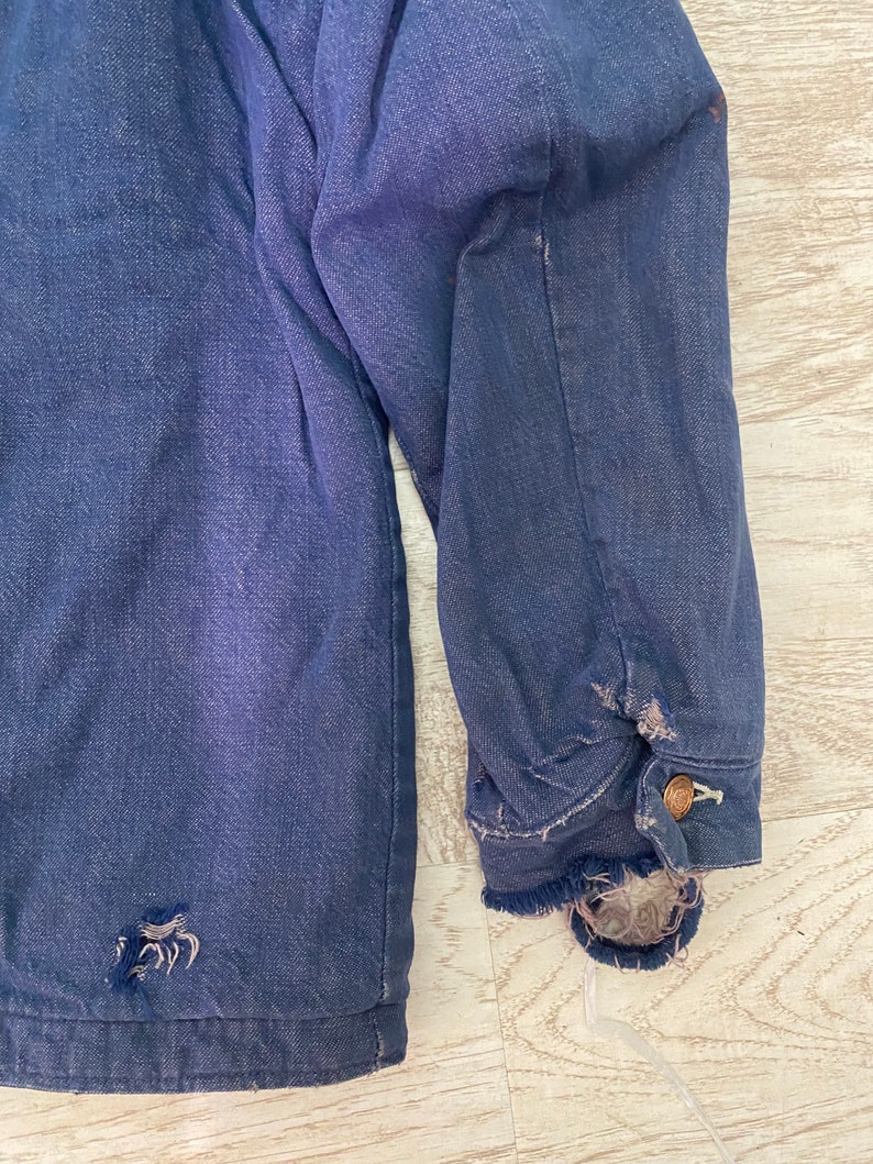May include: A dark blue denim jacket with a frayed hem and a small hole on the left side. The jacket has a button closure and a single button on the right sleeve.