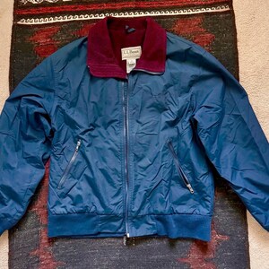 May include: A navy blue L.L.Bean jacket with a burgundy collar and a full zipper. The jacket has two zippered pockets and a ribbed hem and cuffs. The L.L.Bean logo is visible on the collar.