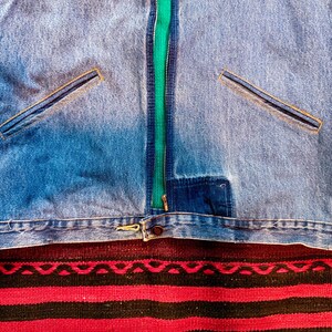 K&ouml;nnte beinhalten: Eine blaue Jeansjacke mit einem gr&uuml;nen Rei&szlig;verschluss und zwei Taschen. Die Jacke liegt auf einem rot-schwarz gestreiften Teppich.