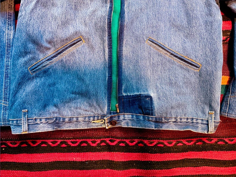 K&ouml;nnte beinhalten: Eine blaue Jeansjacke mit einem gr&uuml;nen Rei&szlig;verschluss und zwei Taschen. Die Jacke liegt auf einem rot-schwarz gestreiften Teppich.