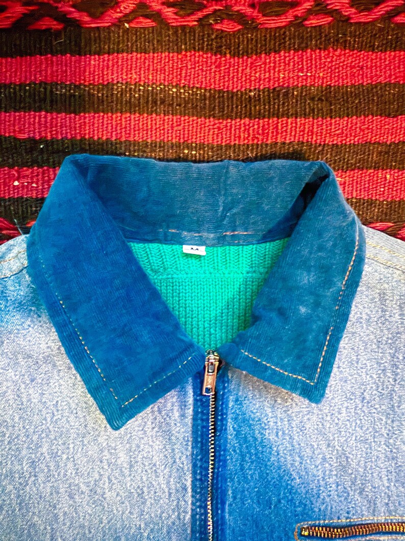 K&ouml;nnte beinhalten: Eine blaue Jeansjacke mit einem blauen Cordkragen und einem gr&uuml;nen Strickfutter. Die Jacke hat einen Rei&szlig;verschluss und eine Tasche auf der linken unteren Seite.