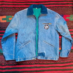 K&ouml;nnte beinhalten: Eine blaue Jeansjacke mit gr&uuml;nem Futter und einem Patch auf der rechten Brustseite. Der Patch zeigt eine Comic-Katze mit dem Text "Cat's Meow".