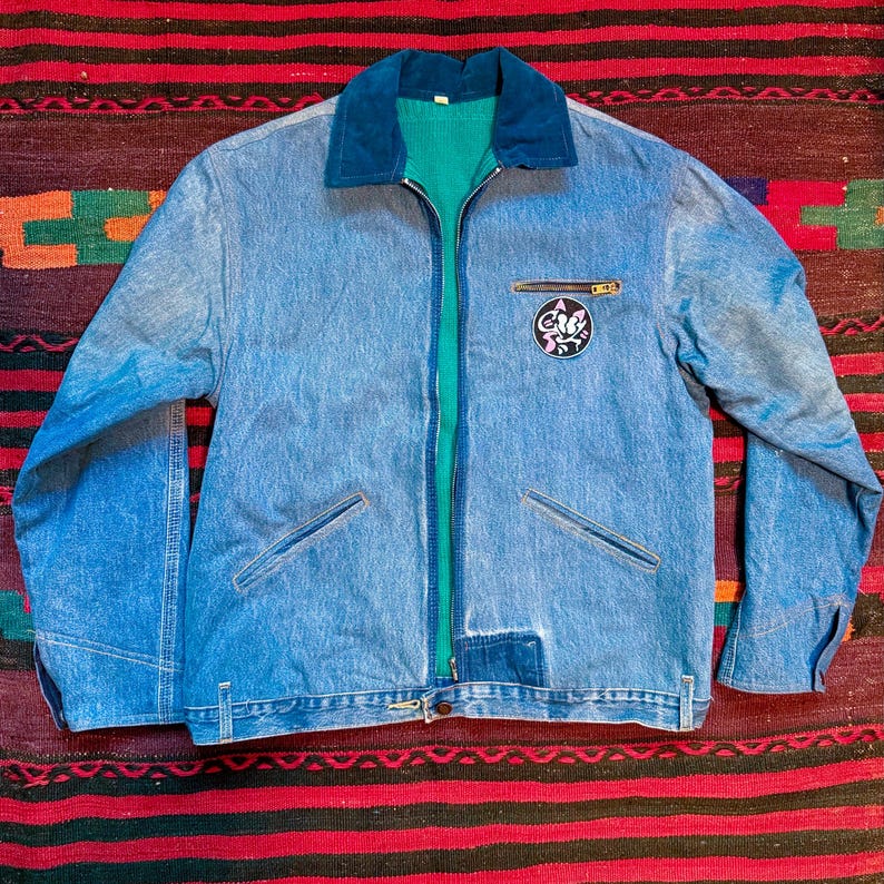 K&ouml;nnte beinhalten: Eine blaue Jeansjacke mit gr&uuml;nem Futter und einem Patch auf der rechten Brustseite. Der Patch zeigt eine Comic-Katze mit dem Text "Cat's Meow".