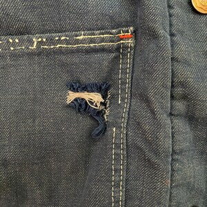 May include: Close-up of a dark blue denim jacket with a white stitched pocket and a small tear in the fabric. The jacket has a gold button on the right side.