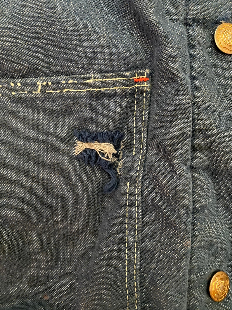 May include: Close-up of a dark blue denim jacket with a white stitched pocket and a small tear in the fabric. The jacket has a gold button on the right side.