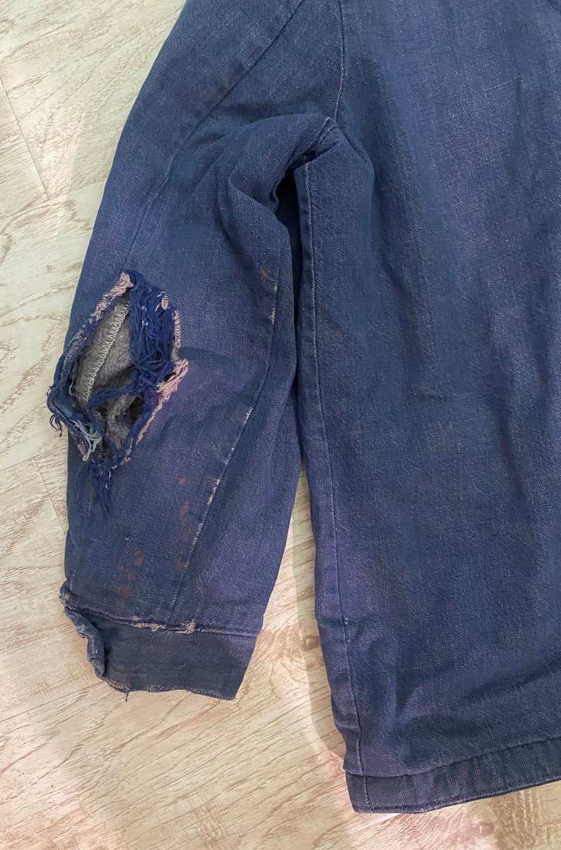 May include: A dark blue denim jacket with a large hole in the elbow. The jacket is worn and has a faded appearance.