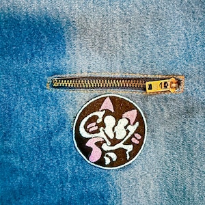 K&ouml;nnte beinhalten: Nahaufnahme einer Jeansjacke mit Rei&szlig;verschluss und einem gestickten Aufn&auml;her in Schwarz und Pink mit einer Comic-Katze.