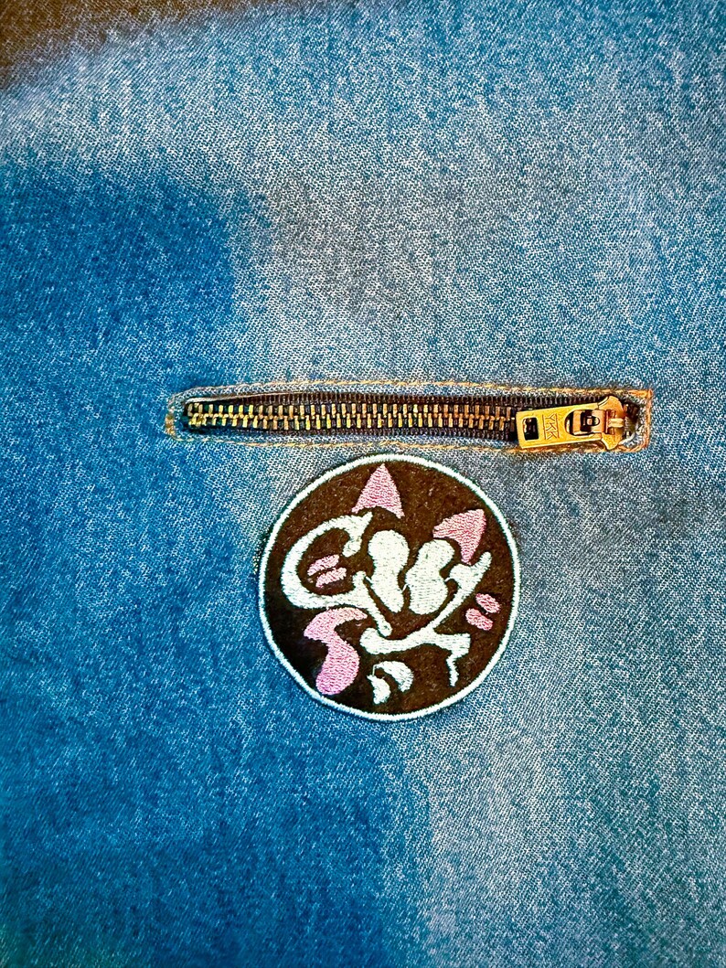 K&ouml;nnte beinhalten: Nahaufnahme einer Jeansjacke mit Rei&szlig;verschluss und einem gestickten Aufn&auml;her in Schwarz und Pink mit einer Comic-Katze.