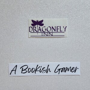 May include: A white background with a purple dragonfly logo that says "Dragonfly Inn" and a white label with the text "A Bookish Gamer".