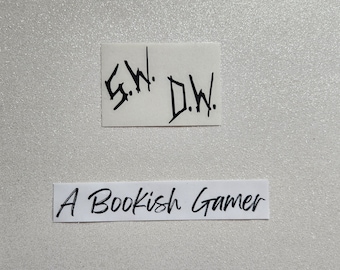 May include: Two white pieces of paper with black handwritten text. The top piece of paper says "S.W. D.W." and the bottom piece of paper says "A Bookish Gamer".