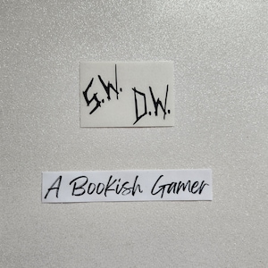 May include: Two white pieces of paper with black handwritten text. The top piece of paper says "S.W. D.W." and the bottom piece of paper says "A Bookish Gamer".