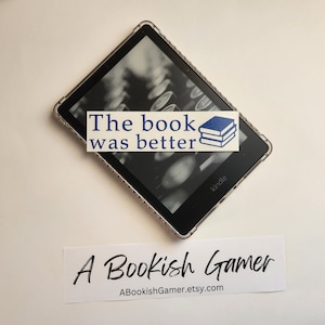 May include: A white sticker with blue text that reads "The book was better" with a stack of three blue books on top of the text. The sticker is placed on a black Kindle e-reader.