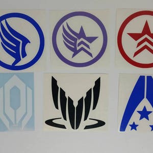 May include: A collection of six stickers featuring various geometric designs and symbols in blue, purple, red, and black. The stickers are on a white background. The designs include stars, wings, and abstract shapes.