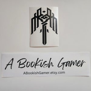 May include: A black and white sticker with a stylized design of a sword and wings. The sticker is on a white background. The text "A Bookish Gamer" and "ABookishGamer.etsy.com" is below the sticker.