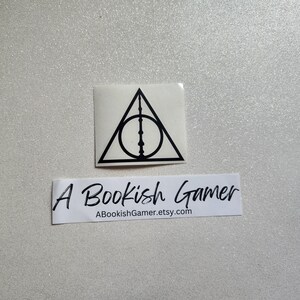 May include: A white sticker featuring the Deathly Hallows symbol in black, a triangle, a circle, and a vertical line. Below is a white rectangle with the text "A Bookish Gamer" and the Etsy shop URL.