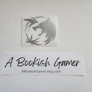 May include: A silver vinyl decal featuring the Witcher logo, a wolf's head with a bird in its mouth, on a white background. The decal is from the Etsy shop ABookishGamer.etsy.com.