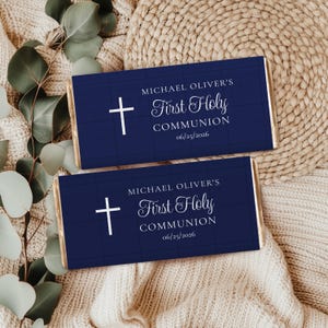 May include: Two navy blue chocolate bar wrappers with gold edges, featuring a white cross and text that reads "MICHAEL OLIVER'S First Holy Communion 06/25/2026". The wrappers are on a cream-colored knitted surface.