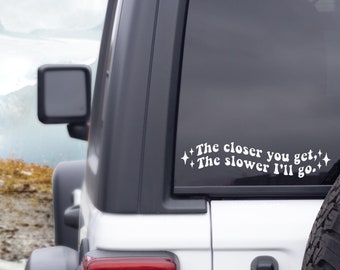 May include: White vinyl decal with the text "The closer you get, The slower I'll go." The decal is displayed on the rear window of a white vehicle. The text is in a stylized font with starburst accents.
