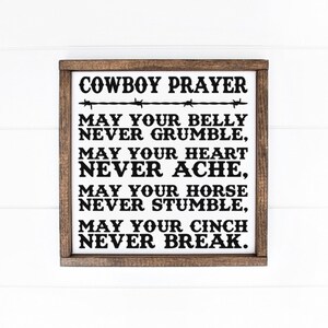 Peut inclure: Un panneau en bois encadr&eacute; avec le texte "COWBOY PRAYER" en noir, accompagn&eacute; d'un motif de fil de fer barbel&eacute;. La pri&egrave;re se poursuit avec les phrases "MAY YOUR BELLY NEVER GRUMBLE", "MAY YOUR HEART NEVER ACHE", "MAY YOUR HORSE NEVER STUMBLE" et "MAY YOUR CINCH NEVER BREAK."