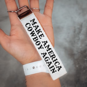 May include: White fabric keychain with black text that reads "Make America Cowboy Again".