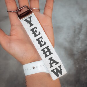 May include: White fabric keychain with the word "YEEHAW" printed in black, attached to a silver metal clasp. The keychain is held up against a gray background.
