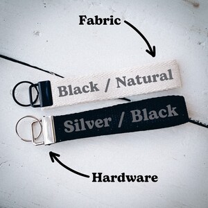 May include: Two keychains with black and silver hardware. The top keychain is made of natural fabric with black text that reads "Black / Natural". The bottom keychain is made of black fabric with silver text that reads "Silver / Black".