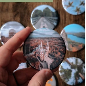 Pu&ograve; includere: Una collezione di bottoni rotondi e colorati con varie immagini stampate su di essi. I bottoni presentano immagini di una chiesa, una strada, un lago e alberi. Il testo "Your Own Photos" &egrave; stampato su uno sfondo verde nella parte superiore dell'immagine.